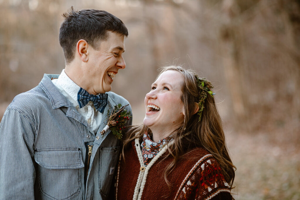 A groom in a pinstripe jumpsuit and bowtie is looking at his bride in a knit poncho. they are both laughing. 