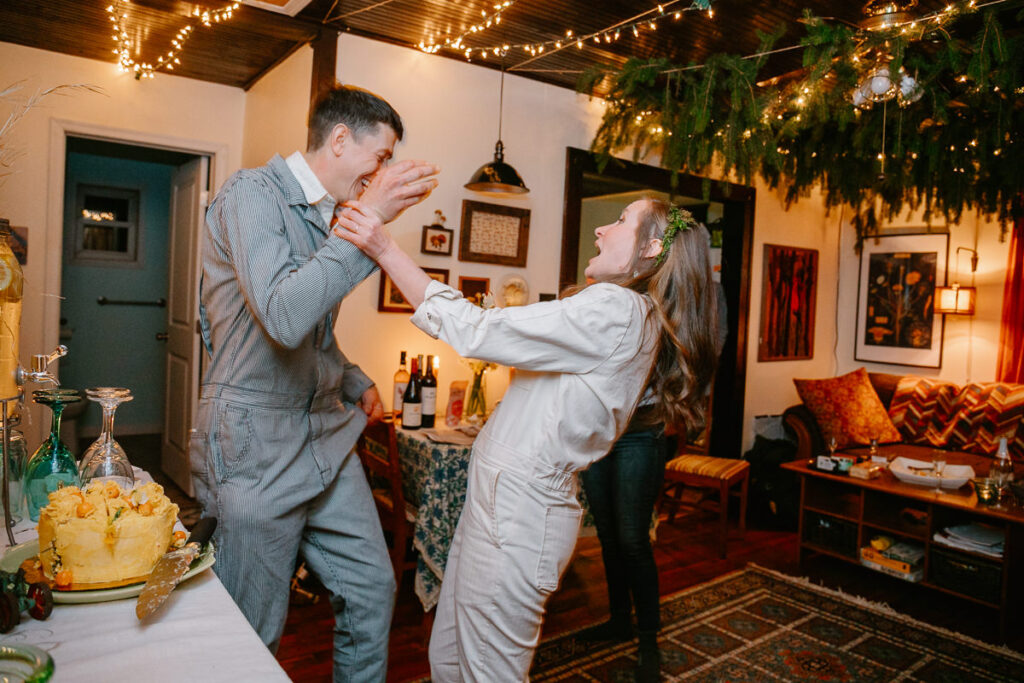 A bride and groom are feeding each other cake during their intimate reception inside their home.  The bride looks afraid that the groom might smash cake in her face. 