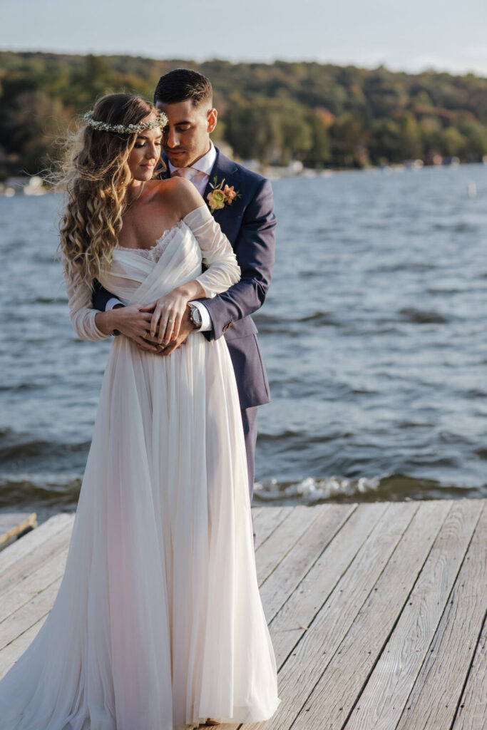 A couple embraces on a dock by Lake Montauk in Sparta New Jersey. The groom has his arms lovingly wrapped around the bride. She is wearing a flower crown and a beautiful boho dress