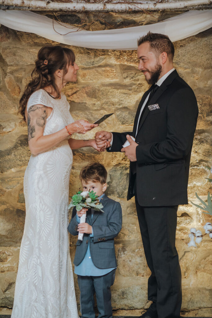 A bride and groom exchanging personal vows. The bride's four year old son is standing in between the couple smelling the brides bouquet.