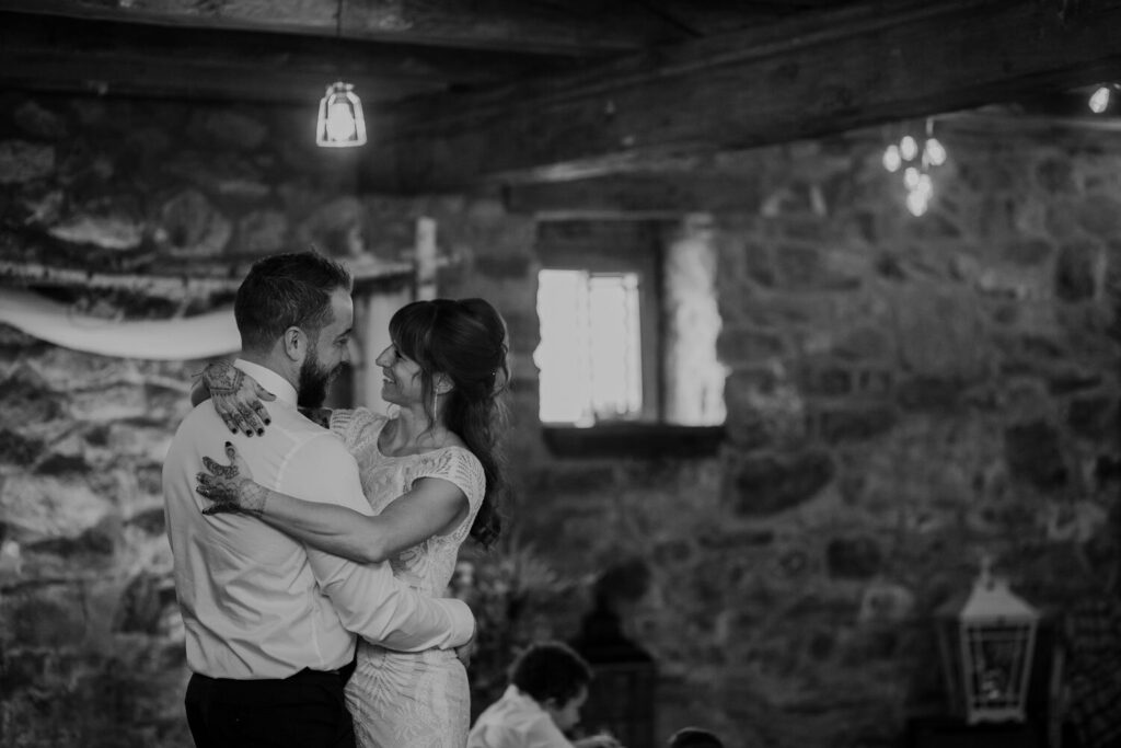 A black and white photo of a bride and groom sharing their first dance Their are twinkling lights, and an old stone wall. The couple is smiling at one another