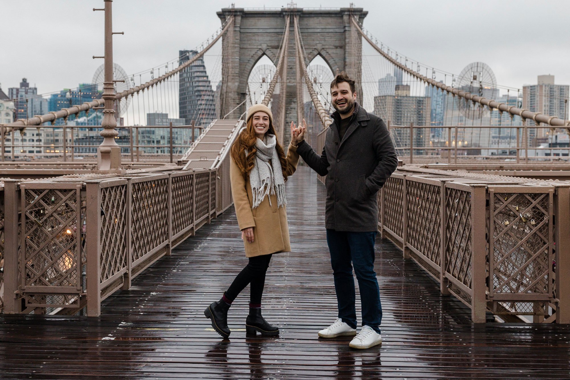 An engaged couple standing on the Brooklyn Bridge saying I love you in sign language