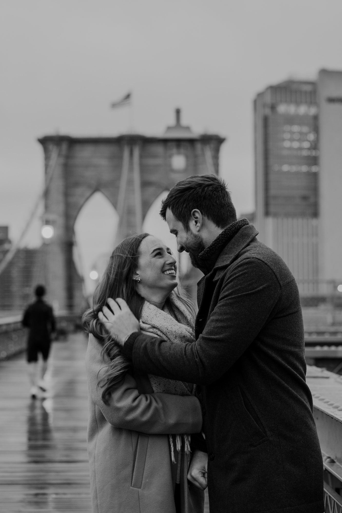 A black and white photo of an engaged couple embracing on the Brooklyn Bridge