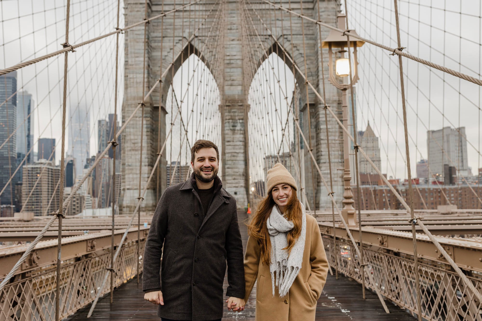 An engaged couple standing while holding hands, on the Brooklyn Bridge
