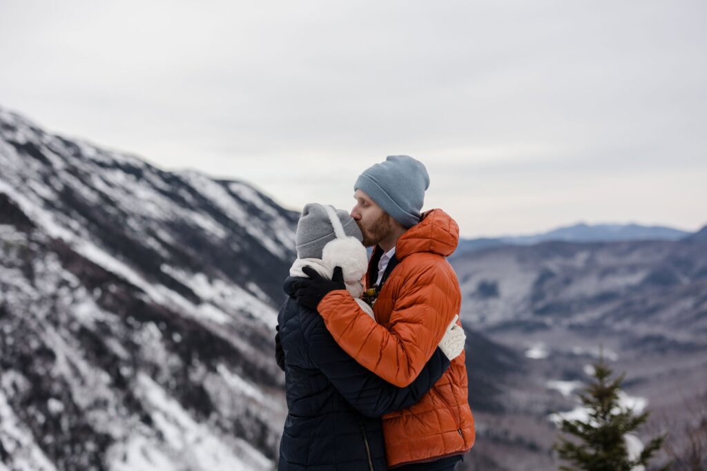 A groom kisses the forehead of his bride during their elopement in the mountains. There are snow covered mountains behind them, and he is wearing a beanie and she is wearing earmuffs