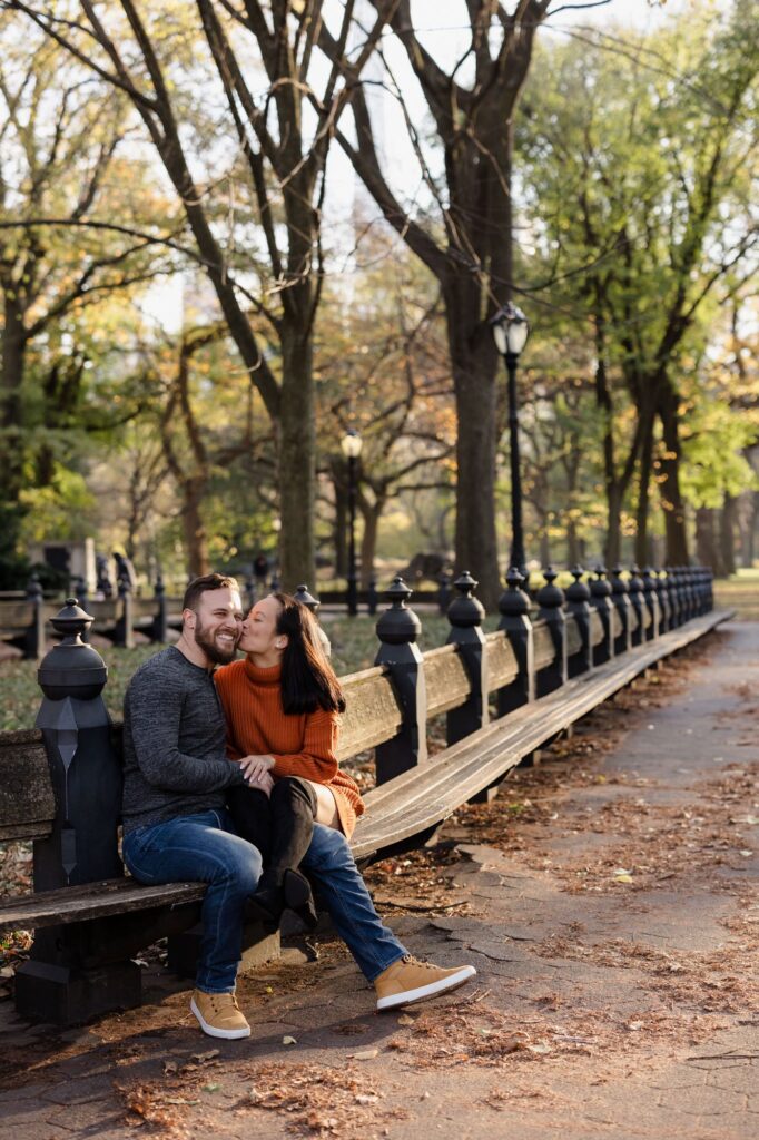 A couple sits on a long bench in central park. She casually has her legs over top of his , and is kissing him on the cheek.