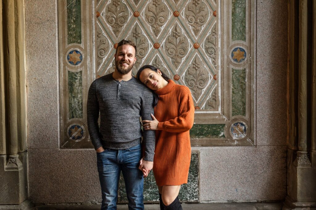 A couple stands in front of a tile wall in Central Park. They are standing next to one another, and she is leaning in with her head on his shoulder.