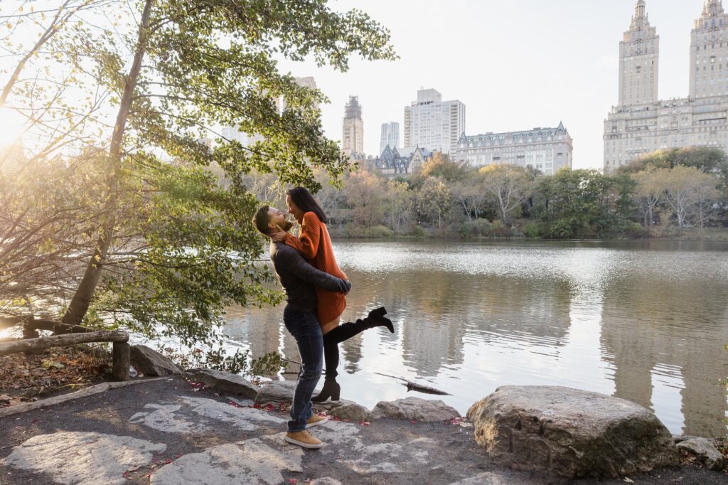 A man romantically picks up his fiancee by the water in Central Park in New York City. 