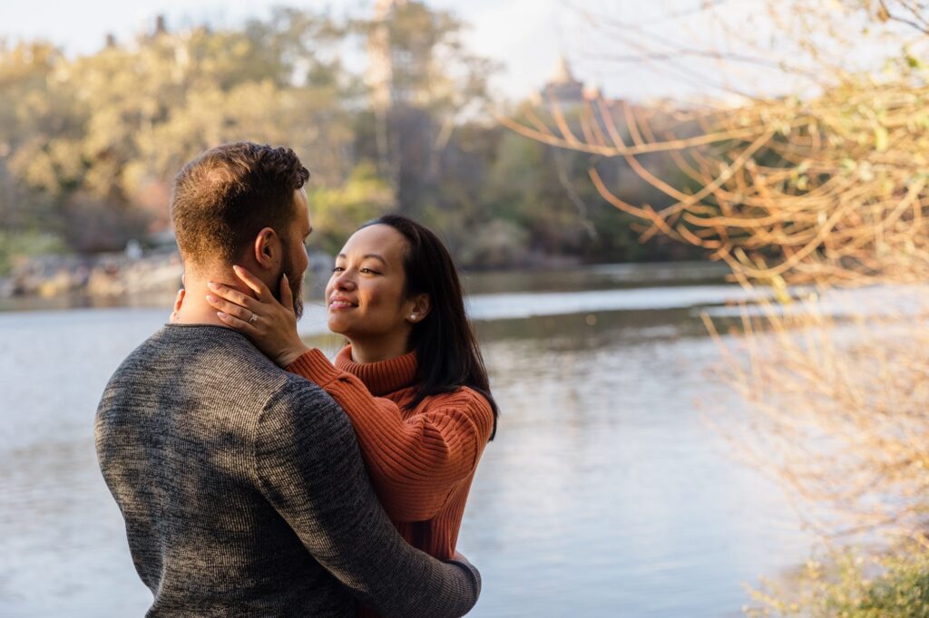 A couple stands by the water during their engagement session. She has her hands gently around his neck, and is gazing at him lovingly