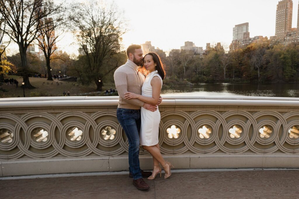 A couple stands on the Bow Bridge in Central Park. The man is leaning into the womans forehead, and she is looking at the camera. You can see the NYC skyline in the background