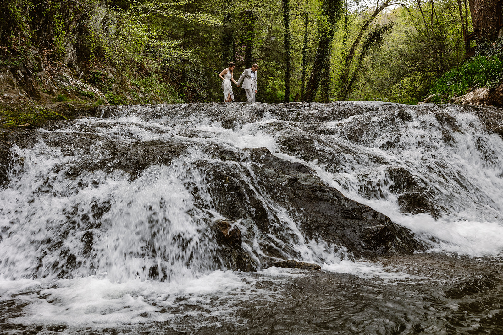 A bride and groom walking in a waterfall during their elopement. The groom is wearing a tan suit with a bowtie, and the bride is wearing a sleeveless dress. the couple is walking across the waterfall holding hands