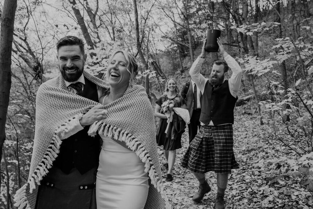 A couple wrapped in a blanket as they hike down a trail after their elopement. They are being followed by their friends. One of them is holding up a speaker playing muisc