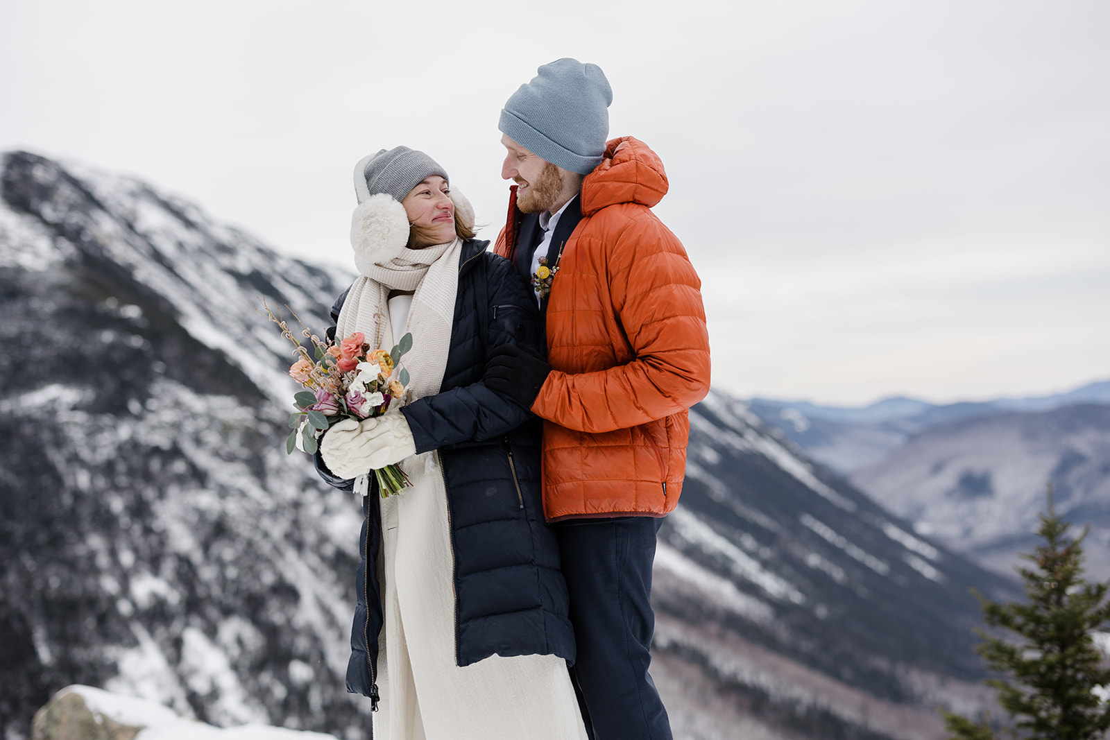 A groom stands behind his bride smiling at her. They are bundled up in warm layers for their winter hiking elopement