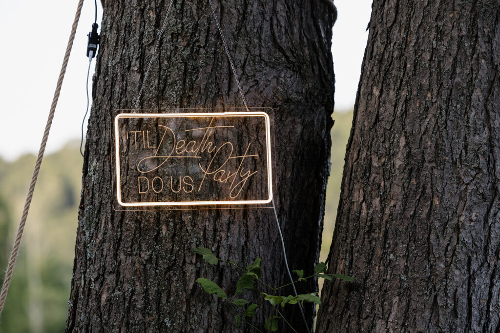 A neon sign hung on a tree that says  "til death do us party"