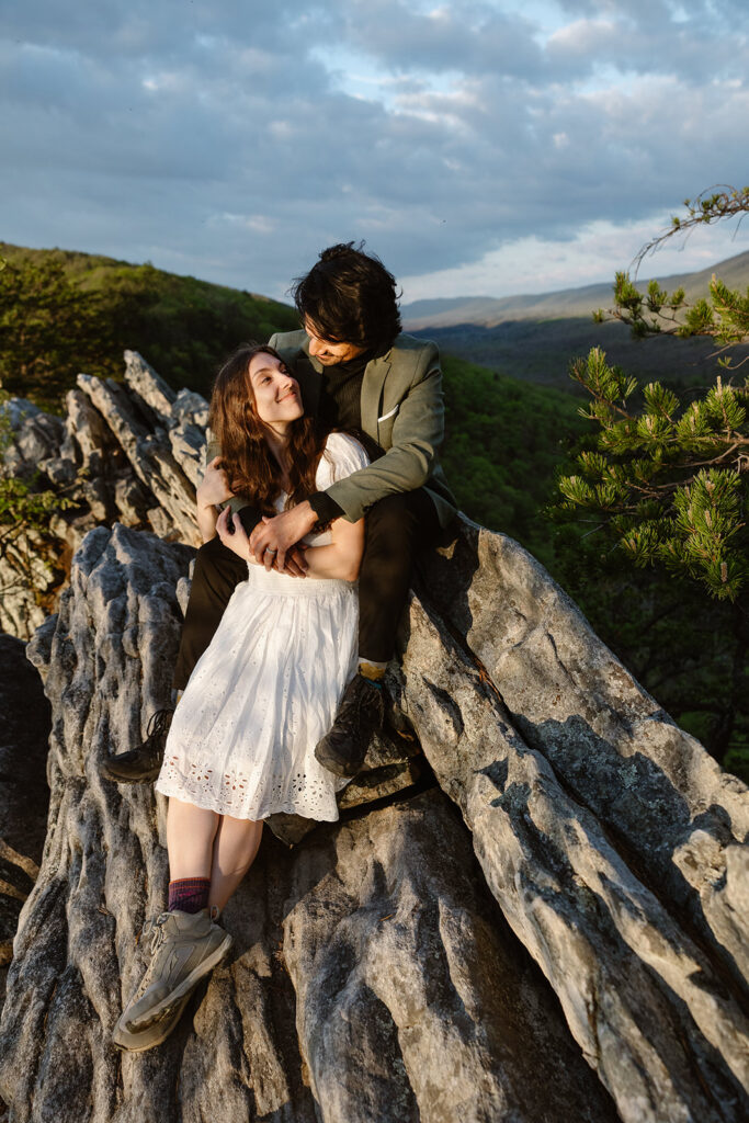 An eloping couple sitting on the top of Buzzard Rock. He is looking down at her lovingly, and she is looking up at him smiling. She is wearing an eyelet dress with her hiking boots, and he is wearing a black turtleneck with a green jacket. They look very happy.