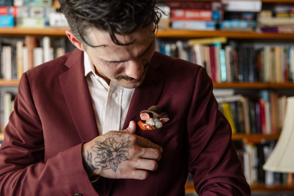 A closeup of the groom and his felted mouse pocket square. The groom has a tattoo on his hand, and is wearing a custom maroon suit. 