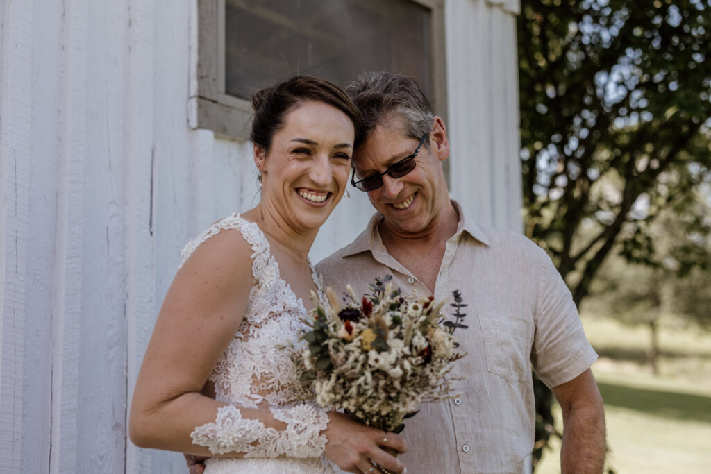 The bride shares a sweet moment with her dad when he sees her for the first time on her elopement day. They are both looking at the camera and smiling. She is wearing an a-line dress with a sheer bodice and sleeves