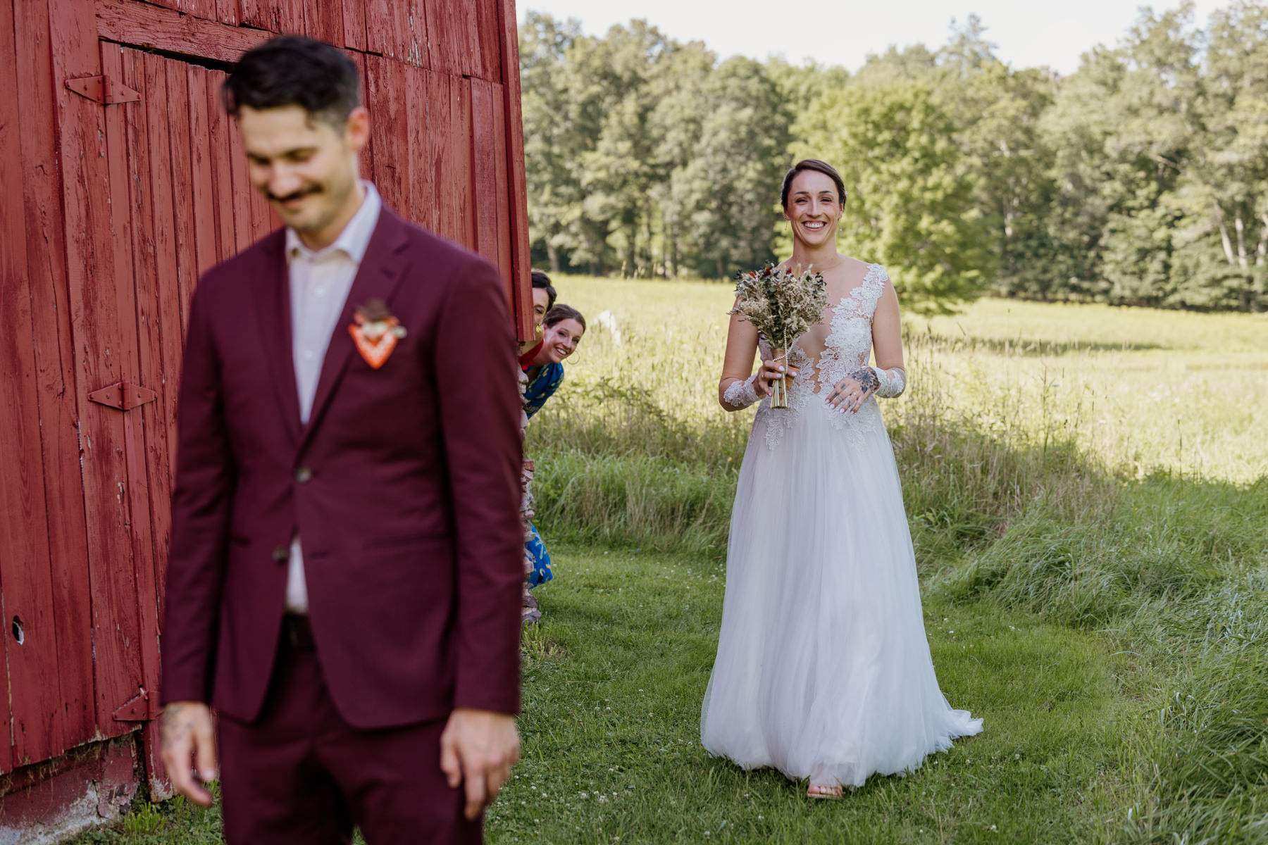 A bride and groom during their first look. They are walking in front of a red barn, and the brides sisters are peeking around the corner to try to get a glimpse. The groom is in the foreground wearing a custom maroon suit, and the bride is wearing an a-line dress with a shear bodice