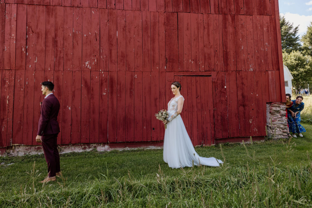 A bride and groom during their first look. They are walking in front of a red barn, and the brides sisters are peeking around the corner to try to get a glimpse. The groom is in the foreground wearing a custom maroon suit, and the bride is wearing an a-line dress with a shear bodice