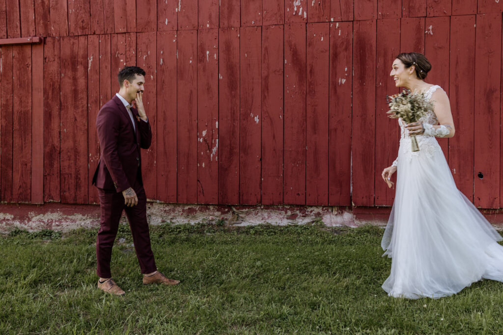 A bride and groom during their first look. The groom has his hand over his mouth with emotion after seeing his bride for the first time. The groom is in the foreground wearing a custom maroon suit, and the bride is wearing an a-line dress with a shear bodice