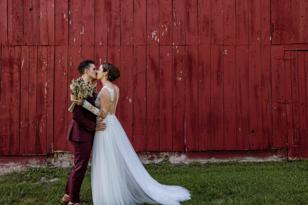 A bride and groom kiss and embrace in front of a red barn during their first look on their elopement day. The groom is wearing a custom maroon suit, and the bride is wearing an a-line dress with a sheer bodice