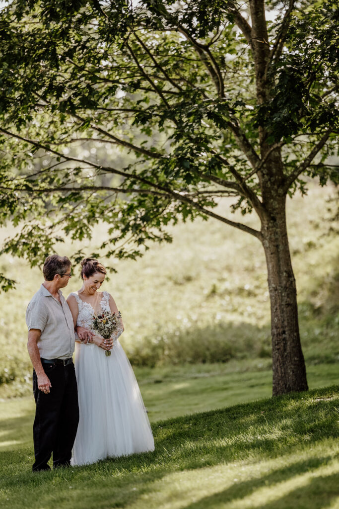 A bride and her dad standing under a tree waiting to walk down the aisle for the brides wedding ceremony. The dad is looking at his daughter, and the bride is laughing. The bride is wearing an a-line dress with a sheer bodice