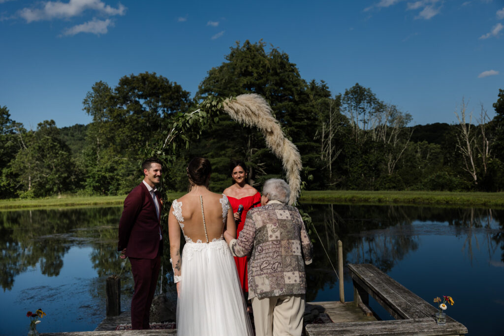 A brides grandma walks her down the aisle towards her groom and the officiant. The ceremony is at a Airbnb in front of a crystal blue pond, and blue skies.