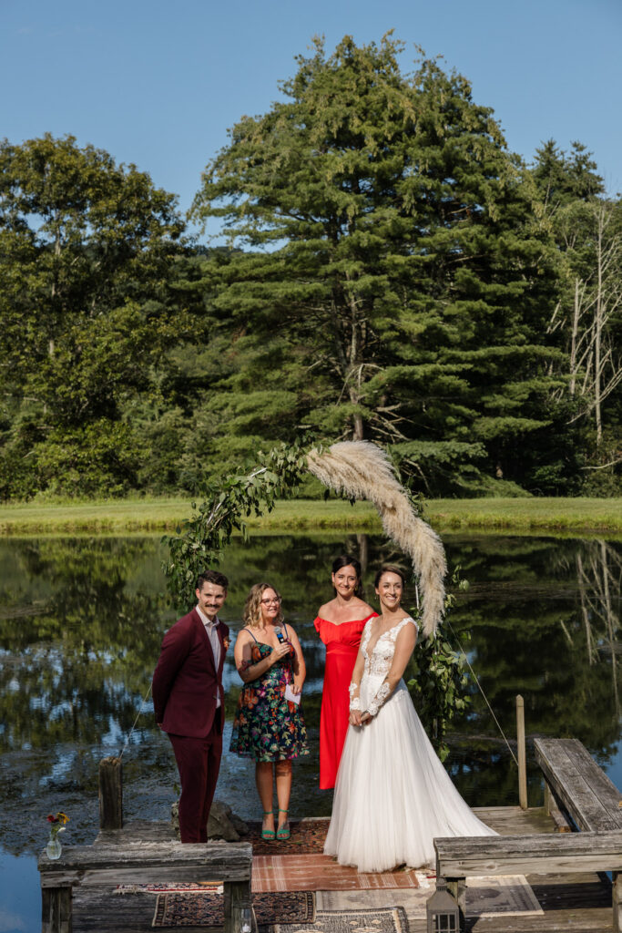 A bride and groom stand on a pier in a pond during their intimate wedding ceremony. They are looking out at their guests. The brides sisters are the officiants. They are standing below a arch made of pampas grass. The groom is wearing a custom maroon suit, and the bride is wearing an a-line dress with a sheer bodice