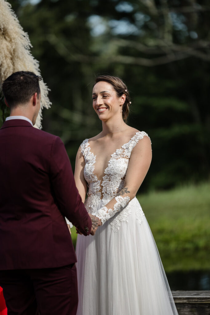 A bride and groom hold hands during their elopement ceremony. Both have tattoos on their hands. The groom is wearing a custom maroon suit, and the bride is wearing an a-line dress with a sheer bodice. The photo is focused on the bride