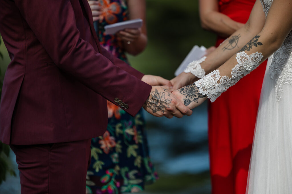 A closeup bride and groom hold hands during their elopement ceremony. Both have tattoos on their hands. The groom is wearing a custom maroon suit, and the bride is wearing an a-line dress with a sheer bodice