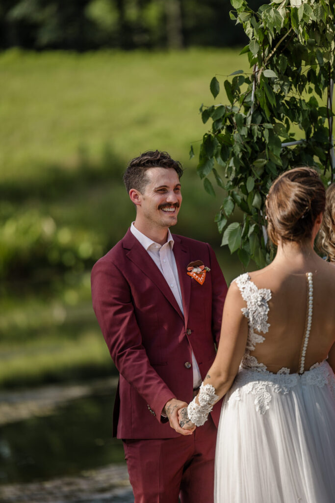 A bride and groom hold hands during their elopement ceremony. Both have tattoos on their hands. The groom is wearing a custom maroon suit, and the bride is wearing an a-line dress with a sheer bodice with buttons going up the back. The photo is focused on the groom