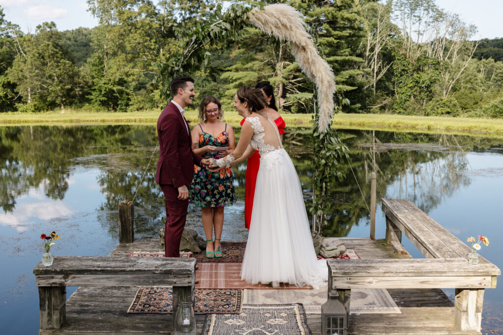 A bride and groom stand on a pier in pond during their intimate ceremony. The bride and groom are holding hands, and the brides sisters are officiating. One is wearing a floral dress and one is wearing a red dress