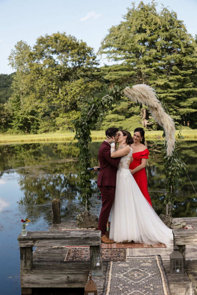 A bride and grooms first kiss during their intimate wedding ceremony.  Both have tattoos on their hands. The bride is holding the grooms face. The groom is wearing a custom maroon suit, and the bride is wearing an a-line dress with a sheer bodice with buttons going up the back. They are standing on a pier in a pond under an arch made of pampas grass.