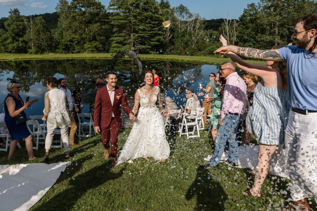 A bride and groom walk up the aisle during their wedding recession as their guests throw biodegradable confetti at them. A bride and groom hold hands during their elopement ceremony. Both have tattoos on their hands. The groom is wearing a custom maroon suit, and the bride is wearing an a-line dress with a sheer bodice