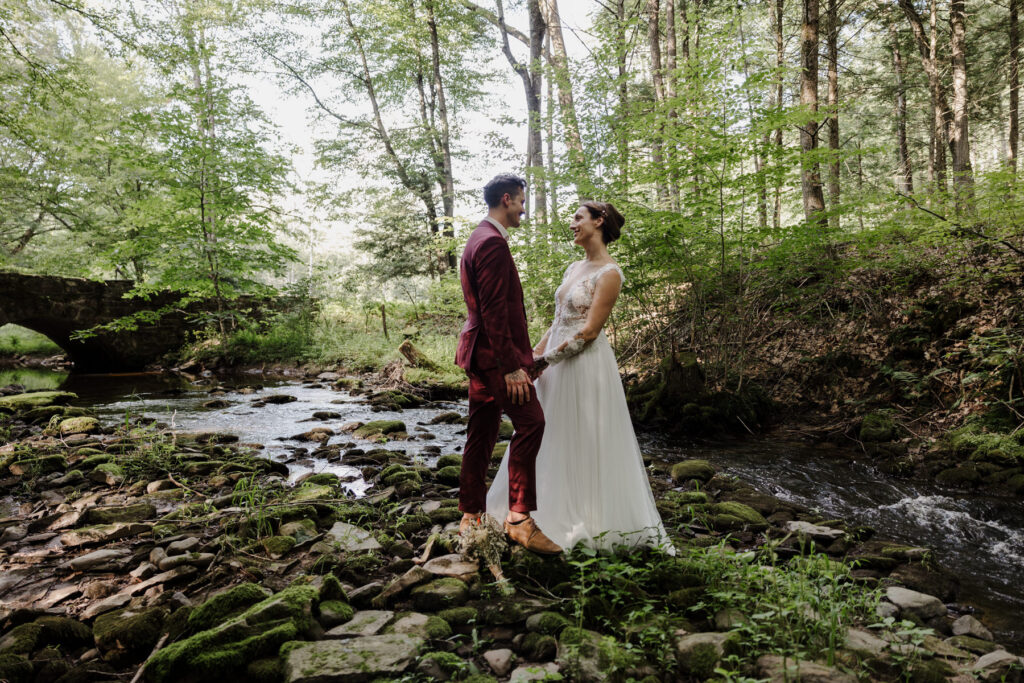 A bride and groom holding hands facing each other on their elopement day. They are standing in a rocky riverbed surrounded by woods. There is a stone bridge in the background. The groom is wearing s custom maroon suit, and the bride is wearing an a-line dress with shear sleeves and bodice.