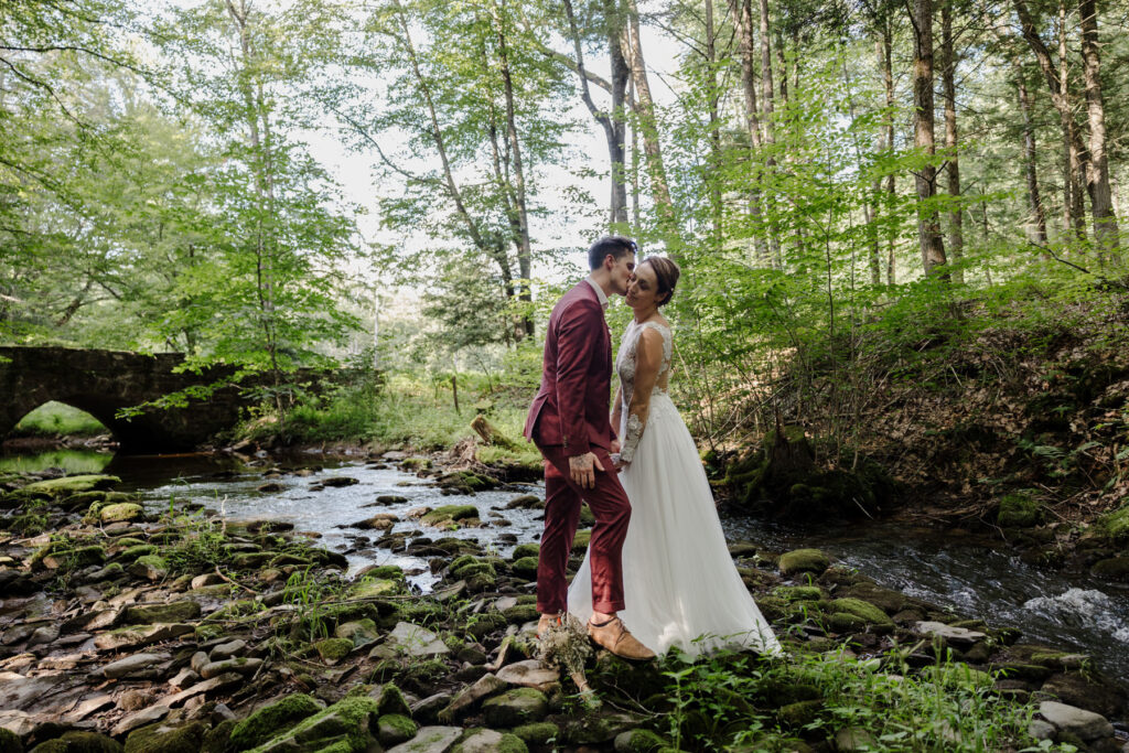 A bride and groom holding hands facing each other on their elopement day. They are standing in a rocky riverbed surrounded by woods. There is a stone bridge in the background. The groom is wearing s custom maroon suit, and the bride is wearing an a-line dress with shear sleeves and bodice.