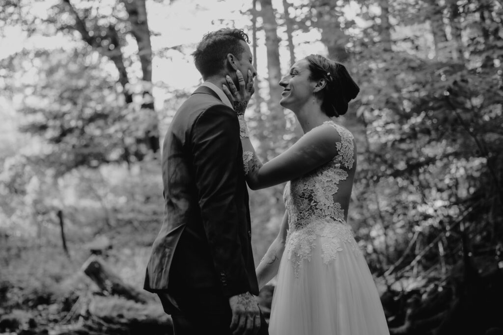 This photo is in black and white. A bride and groom holding hands facing each other on their elopement day. They are standing in a rocky riverbed surrounded by woods. The bride is grabbing the grooms face. The groom is wearing s custom maroon suit, and the bride is wearing an a-line dress with shear sleeves and bodice.