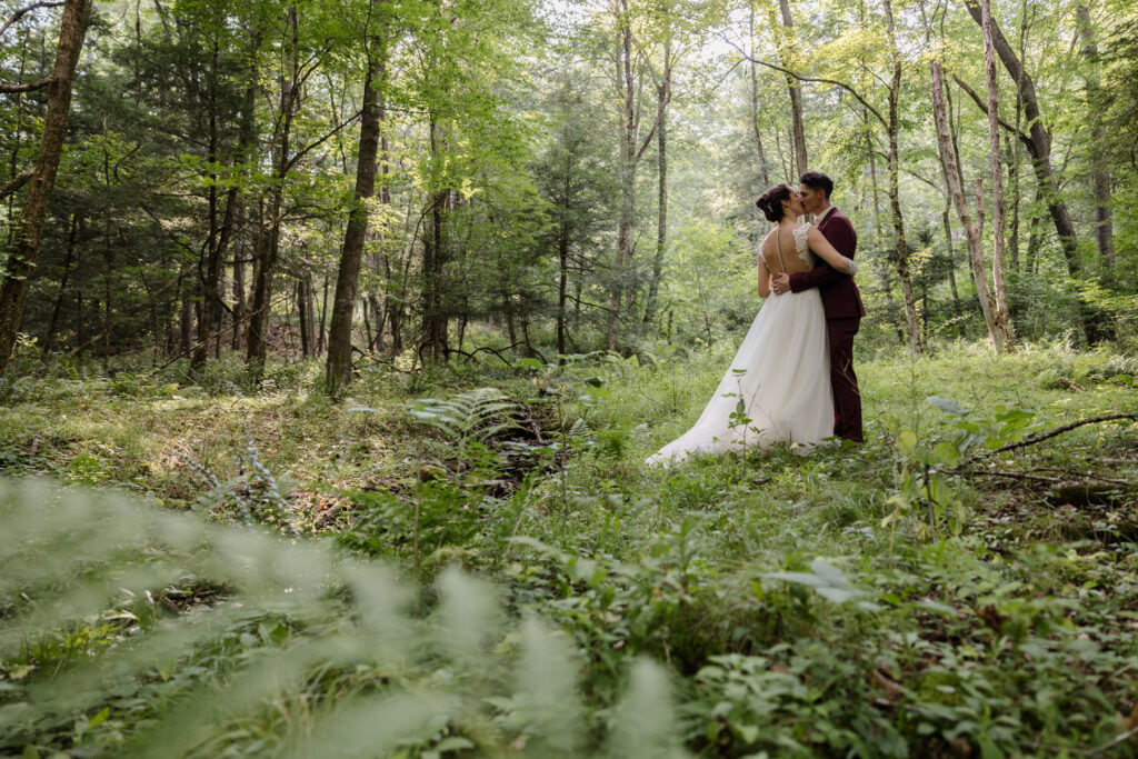 A bride and groom holding hands facing each other on their elopement day. They are standing in a grass surrounded by ferns and woods. There is a stone bridge in the background. The groom is wearing s custom maroon suit, and the bride is wearing an a-line dress with sheer sleeves and bodice with buttons going down the back