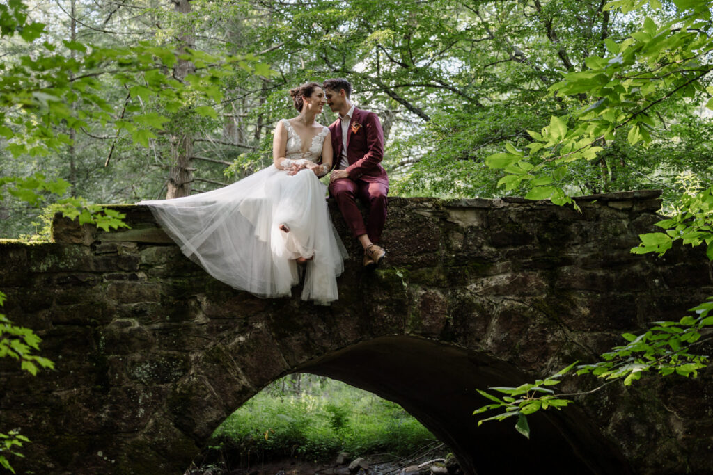 he bride and groom sit together on an arched stone bridge over a stream, with dappled sunlight shining through the surrounding forest, creating a romantic scene.