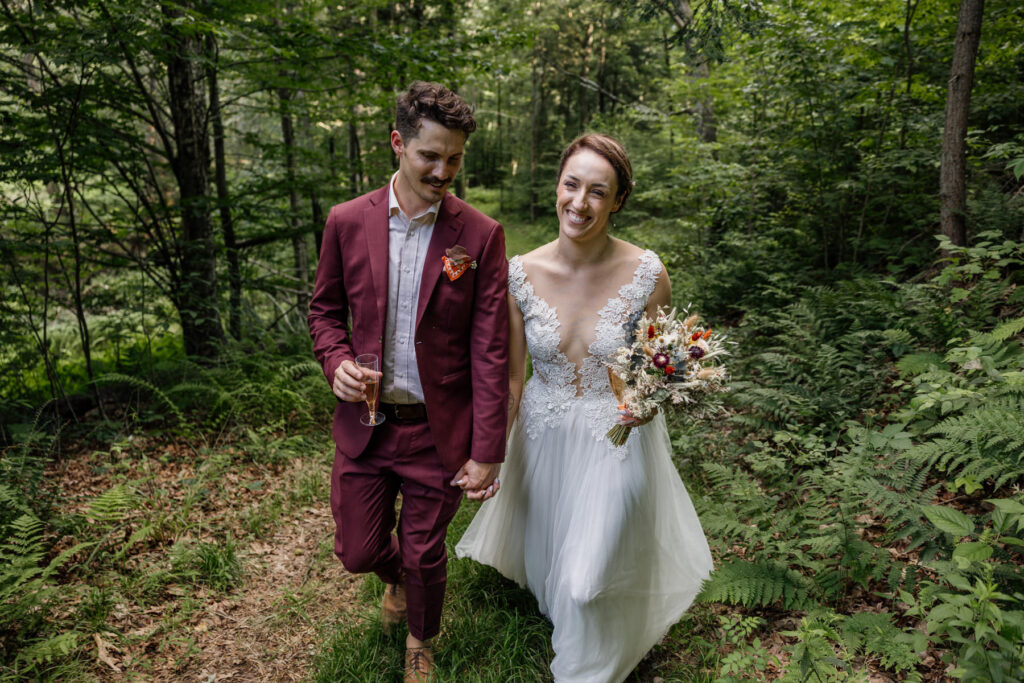 A bride and groom walking hand in hand on their elopement day. The groom is holding a glass of champagne, and the bride is holding her bouquet. They are walking in the woods.