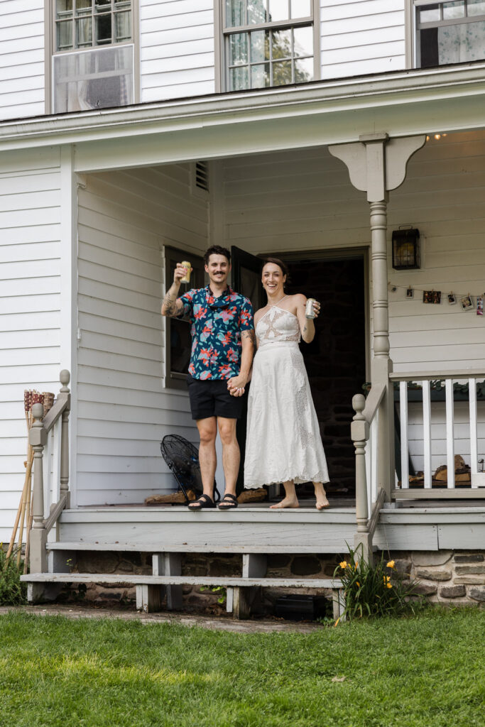 A couple stand on the porch of their airbnb while cheersing beers  The man wears a colorful floral shirt and black shorts, while the woman dons a flowing white dress. 