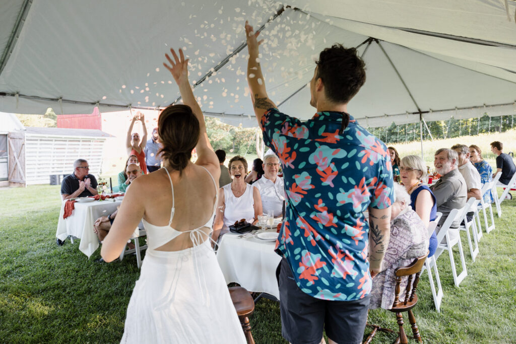 A bride and groom , enter the reception tent of their elopement. The couple is throwing confetti at their friends and family sitting in the tent. The groom is wearing a patterned Hawaiian shirt, and the bride is wearing a cotton dress