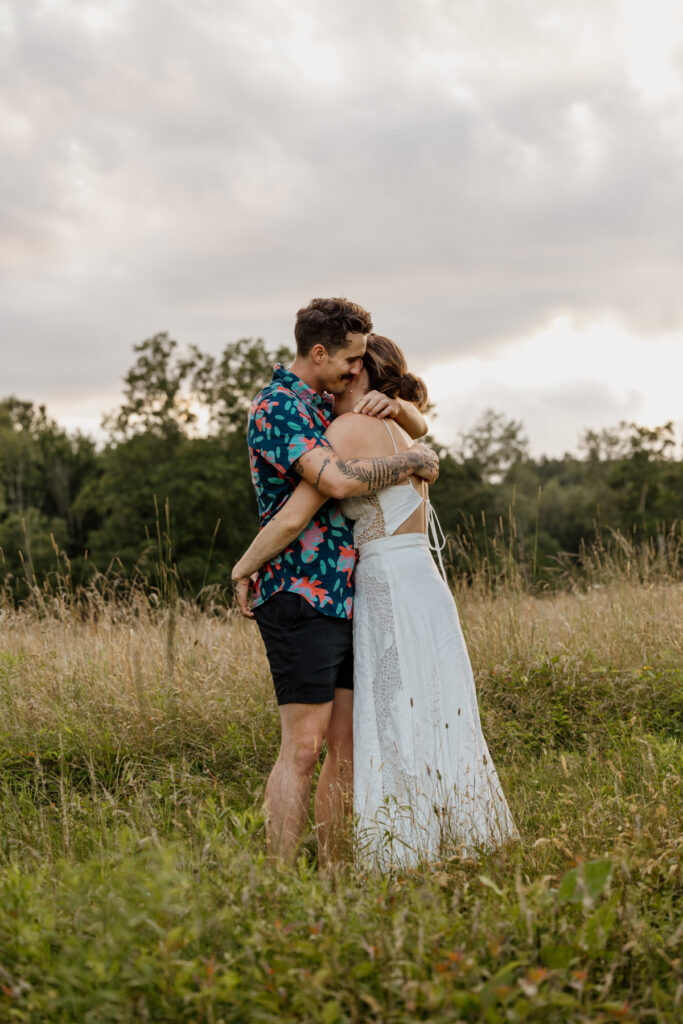 A couple shares an embrace in a sunlit field. The man wears a colorful floral shirt and black shorts, while the woman dons a flowing white dress. The golden light of the sunset creates a romantic atmosphere, illuminating the grassy meadow around them.