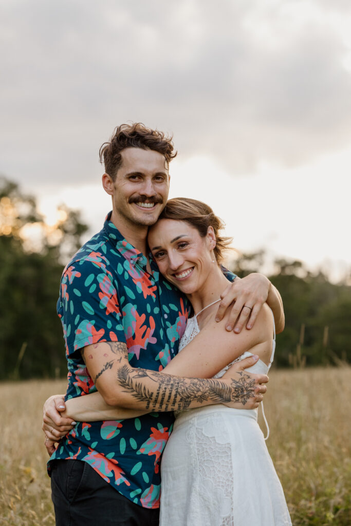 A couple shares an embrace in a sunlit field. They are both looking at the camera and smiling. The man wears a colorful floral shirt and black shorts, while the woman dons a flowing white dress. The golden light of the sunset creates a romantic atmosphere, illuminating the grassy meadow around them.