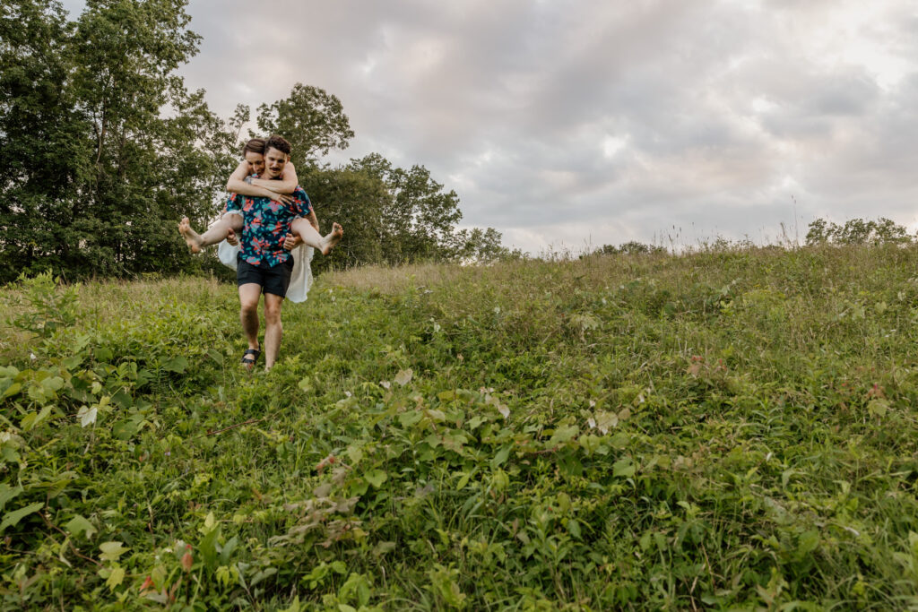 A couple shares a fun picky back ride down a hill. The man wears a colorful floral shirt and black shorts, while the woman dons a flowing white dress, and no shoes.The golden light of the sunset creates a romantic atmosphere, illuminating the grassy meadow around them.