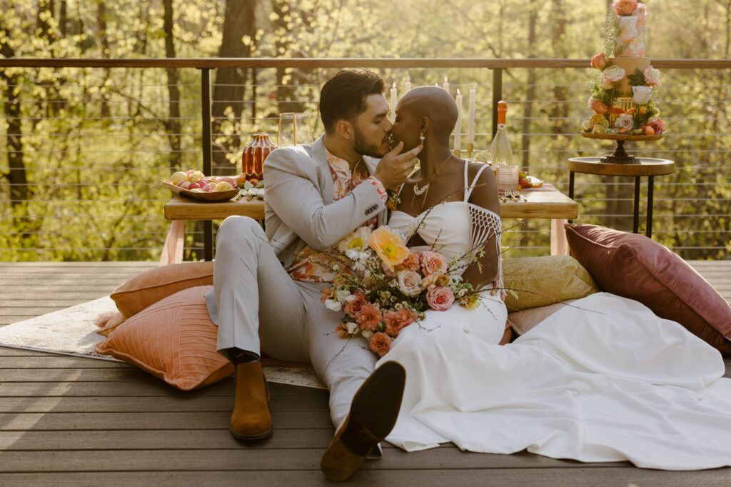 A man and woman are sitting on a deck in front of a luxury picnic on their elopement day. They are kissing, and the groom has his hand on the cheek of the bride