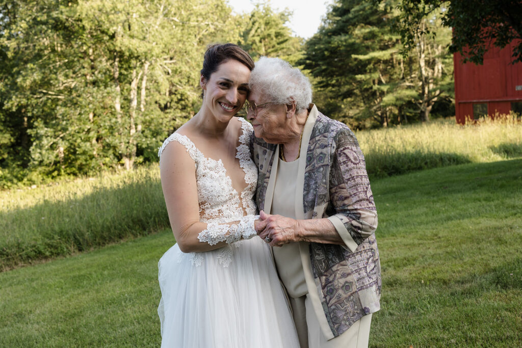 A bride in a lace wedding dress smiles warmly as she embraces an elderly woman with white hair, dressed in a patterned jacket and beige outfit. They hold hands and lean into each other affectionately, surrounded by a serene green landscape.