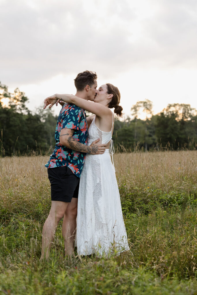 A couple shares a tender kiss in a sunlit field. The man wears a colorful floral shirt and black shorts, while the woman dons a flowing white dress. The golden light of the sunset creates a romantic atmosphere, illuminating the grassy meadow around them.