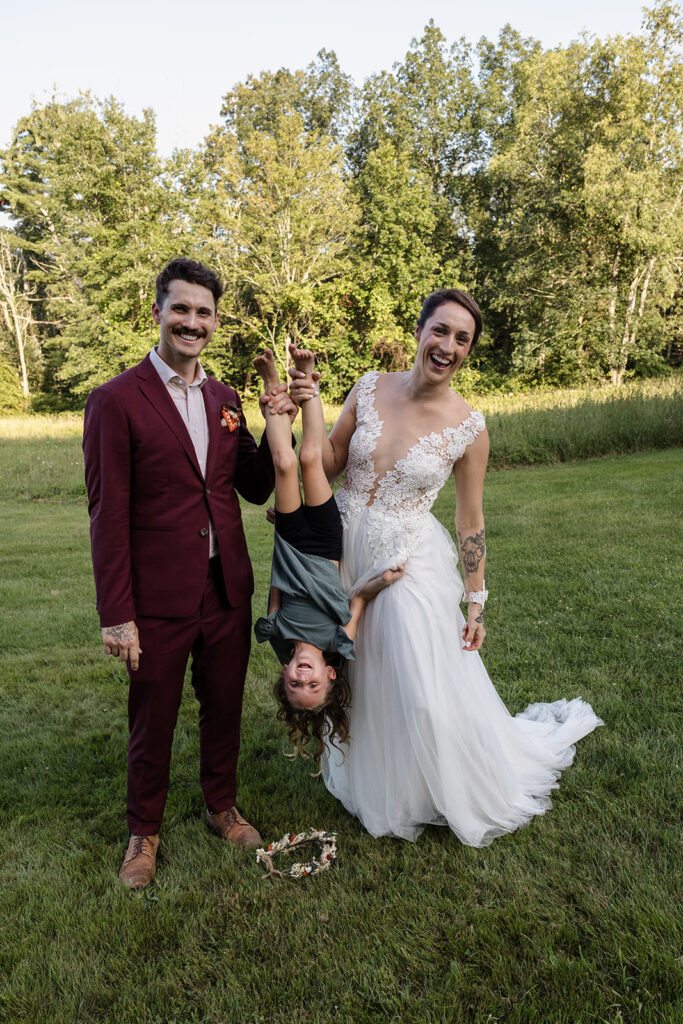 The bride and groom pose playfully with a young girl held upside down, smiling widely in a grassy field during their wedding celebration.