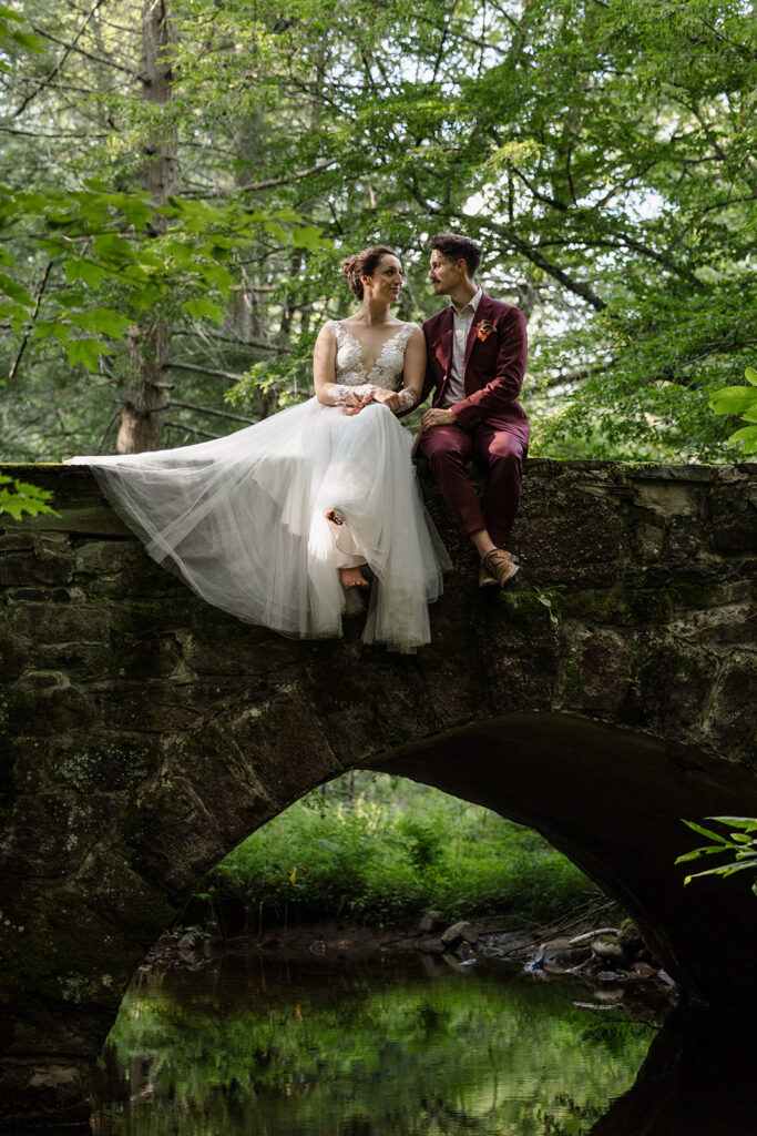 The bride and groom sit together on an arched stone bridge over a stream, with dappled sunlight shining through the surrounding forest, creating a romantic scene.